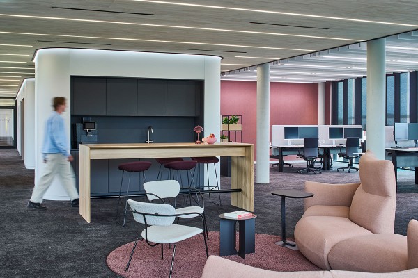 Die offene Bürofläche bei Arethia kombiniert funktionale Arbeitsplätze mit wohnlichen Zonen. Im Vordergrund lädt eine Lounge mit rosafarbenem Sofa und Sesseln zum Austausch ein. Dahinter befinden sich eine Küchenzeile und ein hoher Tresen mit Barhockern.
