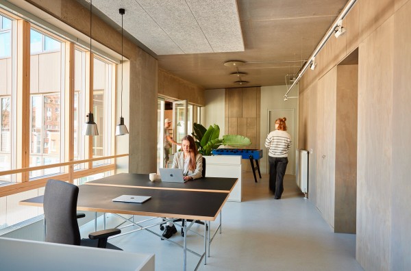 Das helle Büro bei Karsten Jahnke kombiniert Sichtbetondecken mit warmen Holzwänden und einer großen Fensterfront. Eine Mitarbeiterin arbeitet fokussiert an einem der schwarzen Tische mit Laptop. Im Hintergrund befinden sich eine große Zimmerpflanze und ein blauer Kicker.