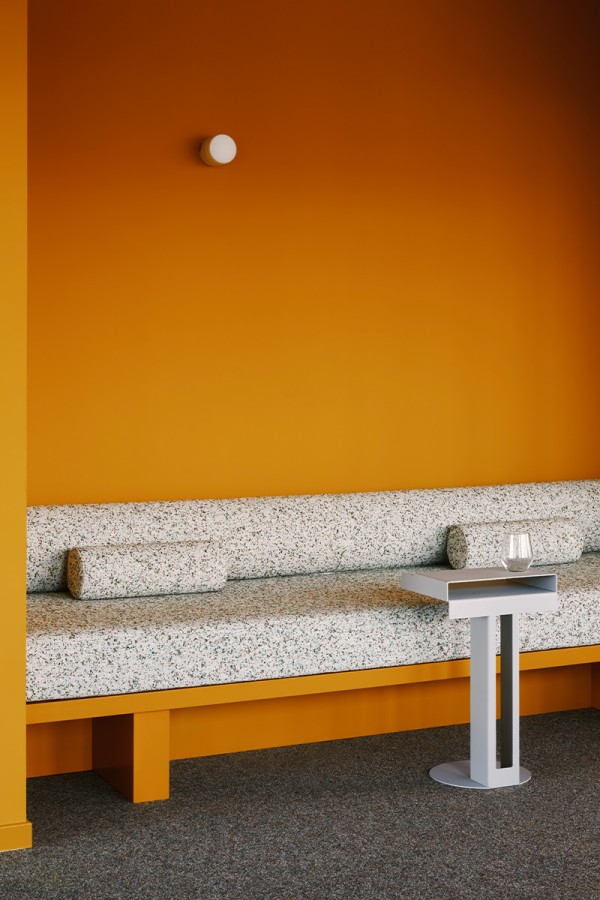Ein moderner Aufenthaltsbereich bei ng-voice besticht durch eine kräftig orangefarbene Wand und eine passende Holzbank. Die lange Sitzfläche ist mit gemustertem Stoff bezogen, davor steht ein minimalistischer grauer Beistelltisch.