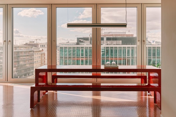 Der lichtdurchflutete Raum bei ng-voice besticht durch ein modernes Set aus einem langen Tisch und zwei Bänken in glänzendem Rot. Eine minimalistische Pendelleuchte hängt über dem Ensemble. Dahinter bietet eine breite Fensterfront Aussicht auf urbane Gebäude.