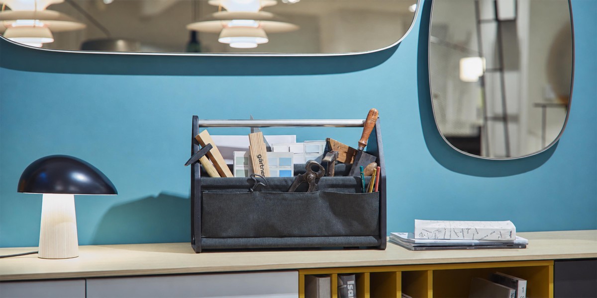 Im gärtner Showroom steht eine dunkle Werkzeugtasche gefüllt mit Werkzeugen und Farbkarten auf einer hellen Konsole. Daneben befinden sich eine kleine Tischleuchte und Bücher vor einer blauen Wand mit Spiegel.