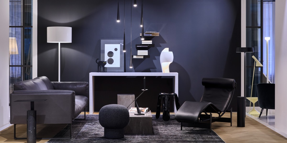 Im gärtner Showroom zeigt sich eine edle Lounge mit schwarzem Ledersofa, passender Chaiselongue und Hocker vor dunkler Wand. Filigrane Pendelleuchten, Stehlampen und eine weiß geflieste Konsole setzen stilvolle Lichtakzente.