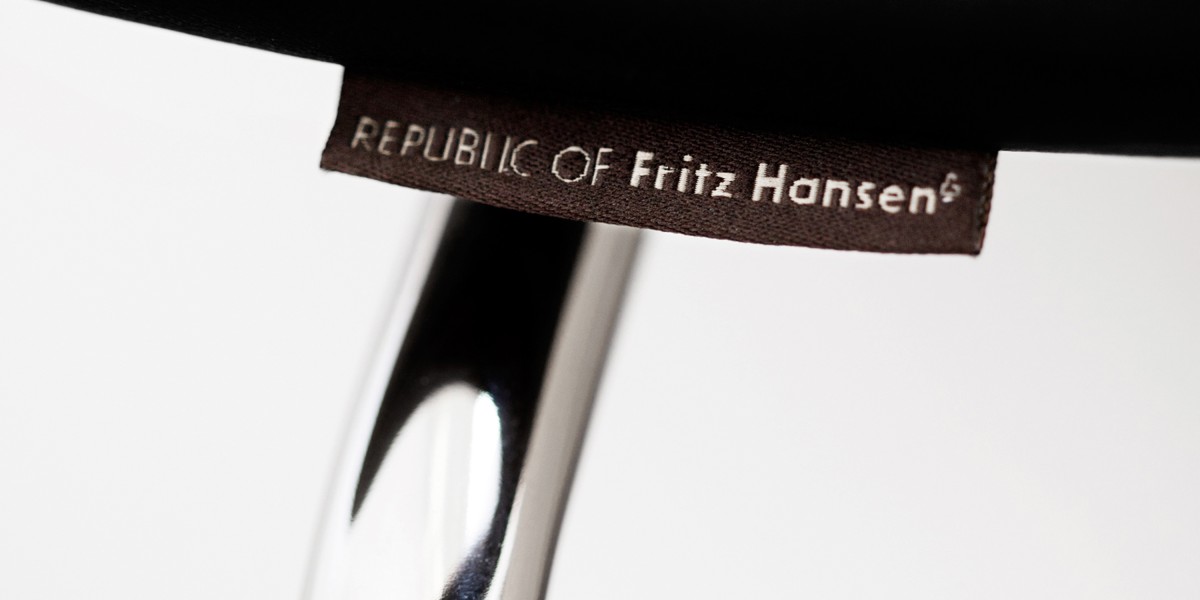 Die Nahaufnahme zeigt ein braunes Stoff-Etikett mit der hellen Aufschrift „Republic of Fritz Hansen“. Das Label ist an der Unterseite des dunklen Sitzpolsters oberhalb des verchromten Metallgestells angebracht.