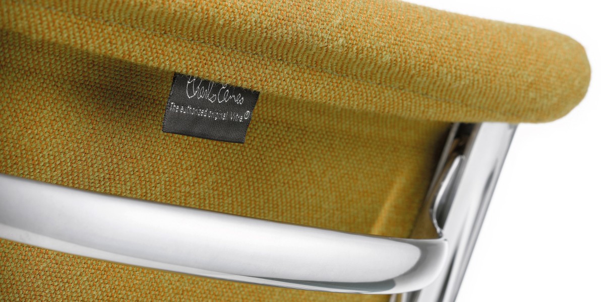Die Nahaufnahme zeigt die Rückenlehne eines Eames-Klassikers von Vitra mit senfgelbem Stoffbezug und verchromtem Metallrahmen. Ein angenähtes schwarzes Stoffetikett bestätigt die Echtheit als lizenziertes Original.